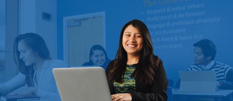 A smiling woman typing on her laptop in a busy open workspace